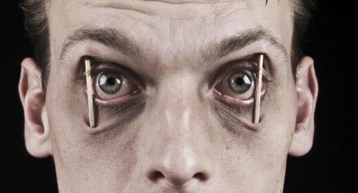 A close-up image of a person's face with clothespins holding open their upper and lower eyelids, preventing them from closing their eyes.