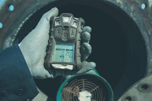 Hand in a protective glove holding a portable gas detector displaying readings for oxygen and hazardous gases.