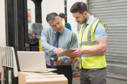 Supervisor demonstrating safety leadership by discussing a checklist with a worker in a warehouse, both wearing safety gear, with a laptop and packages in the background.