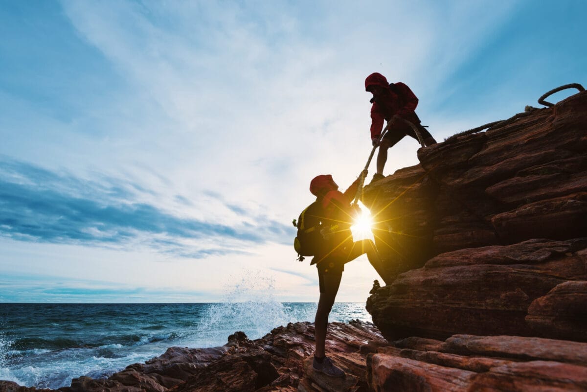 Two climbers practising supervisor safety leadership, with one climber helping another up a rocky cliff by pulling on a rope, set against a scenic ocean backdrop at sunset.