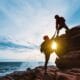 Two climbers practising supervisor safety leadership, with one climber helping another up a rocky cliff by pulling on a rope, set against a scenic ocean backdrop at sunset.